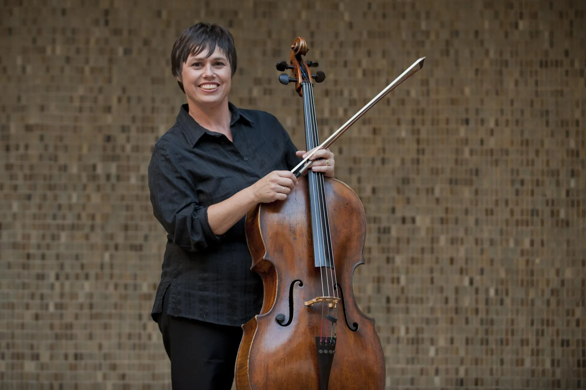 Anmari standing in front of a brown brick wall, holding her bow and cello. She is wearing a black shirt.
