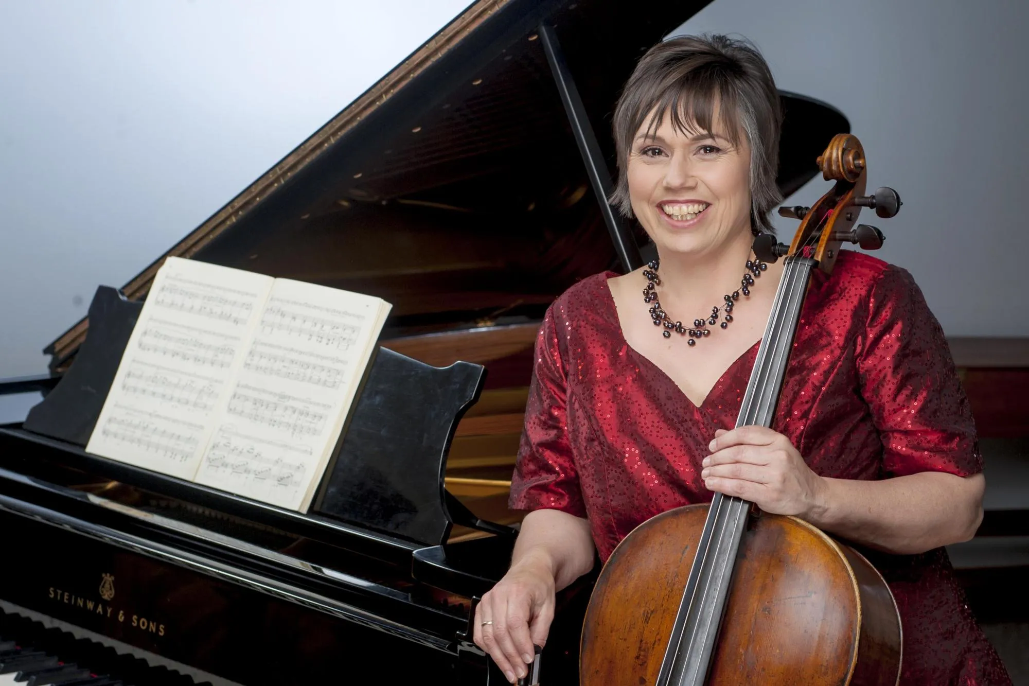 Anmari holding her cello and leaning against a grand piano, wearing a dark red sequined blouse. A volume of sheet music is on the music stand, and the piano's lid is propped open.