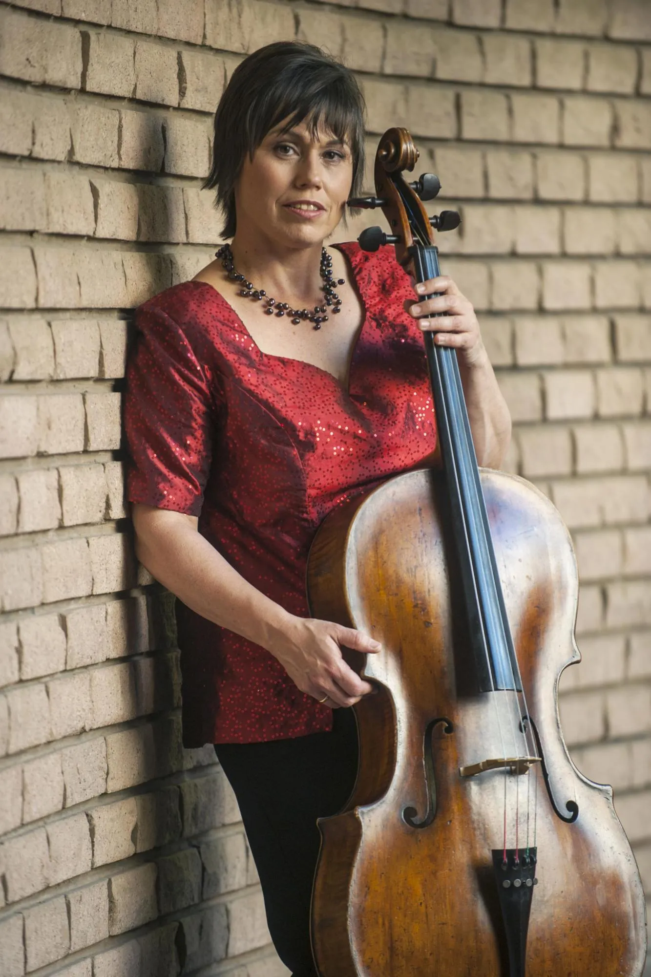 Anmari holding her cello, leaning against a brick wall which recedes into the background. She is wearing a dark red sequined blouse.