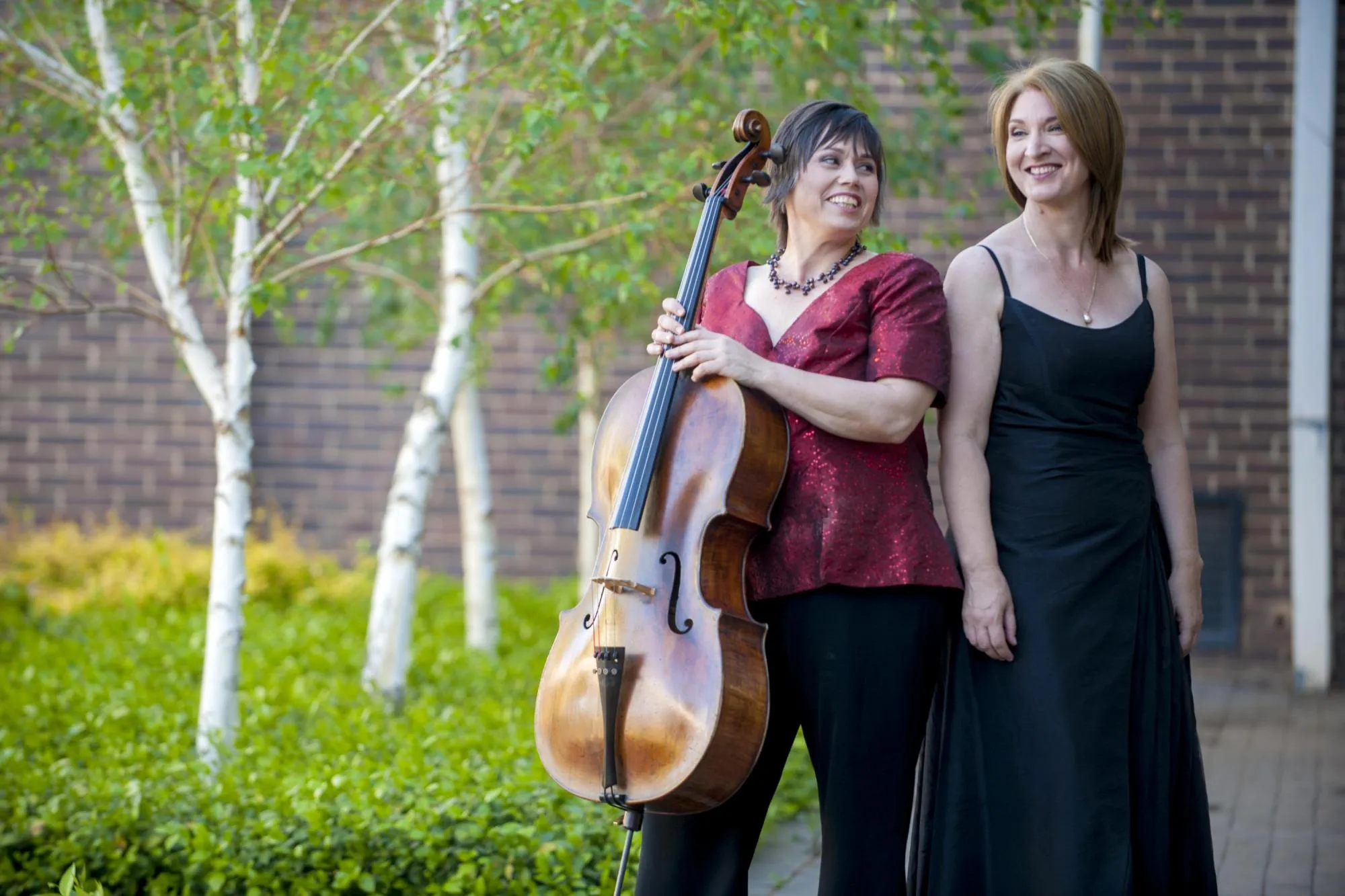 Anmari and Ilse standing against each other in a courtyard of green bushes and trees with white bark, both smiling. Anmari is holding her cello. She is wearing a dark red sequined blouse, and Ilse a black dress.