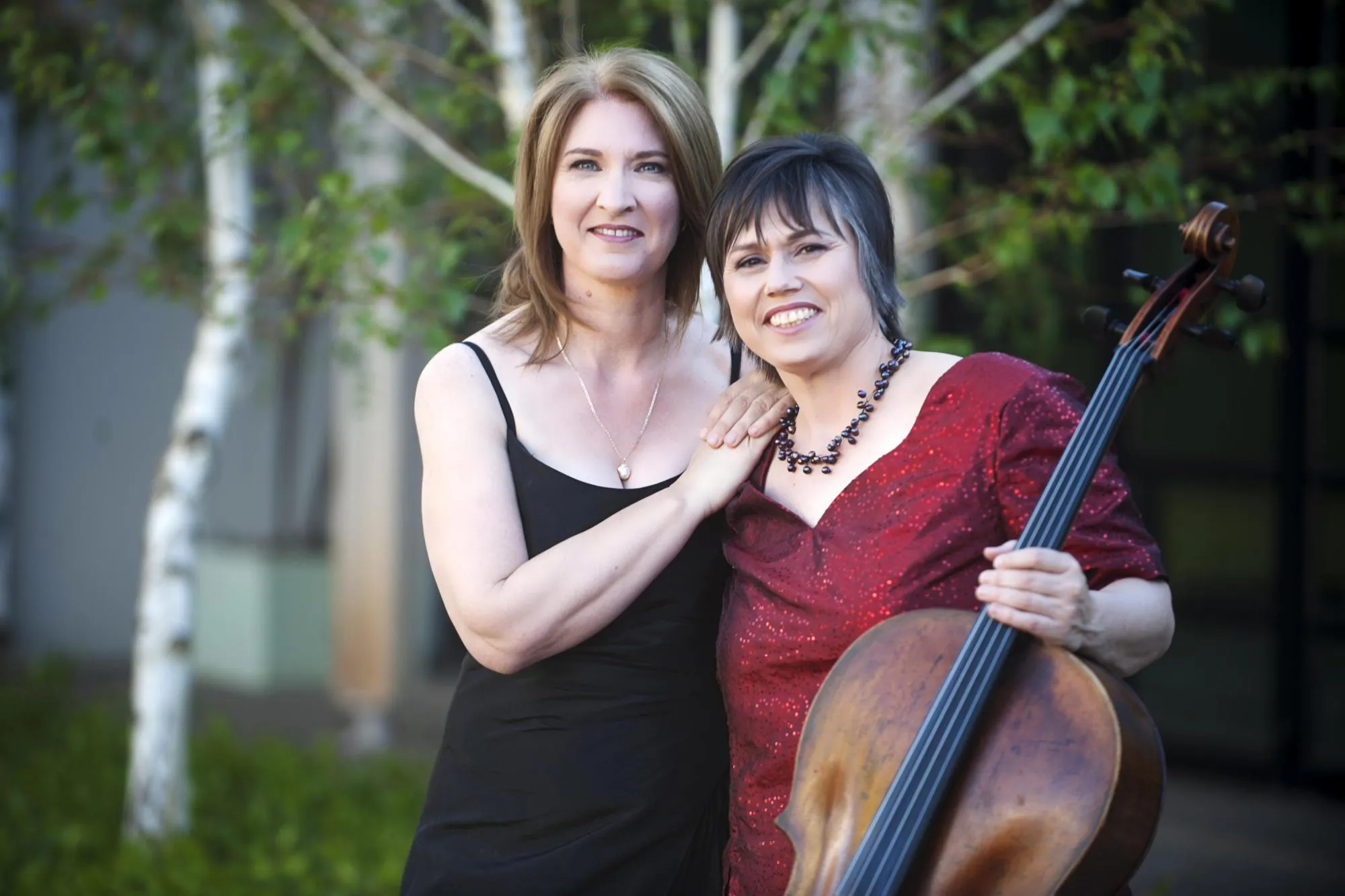 Anmari and Ilse next to each other in a courtyard. Anmari is holding her cello and in the background are trees with white bark. Anmari is wearing a dark red sequined blouse, and Ilse a dark dress.