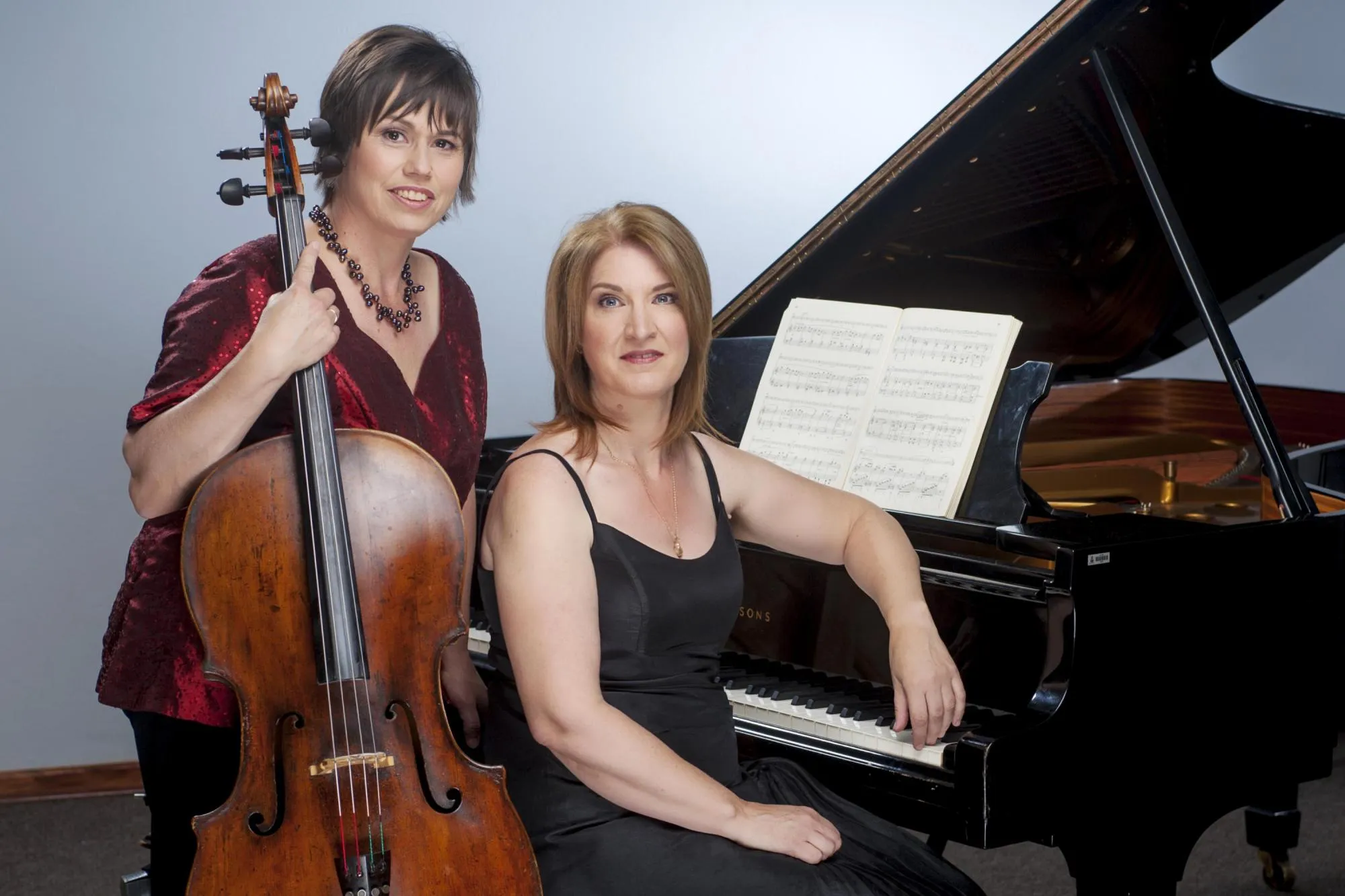 Anmari holding her cello, standing next to Ilse, who is sitting at a grand piano. The piano lid is propped open. Anmari is wearing a dark red sequined blouse, and Ilse a black dress.