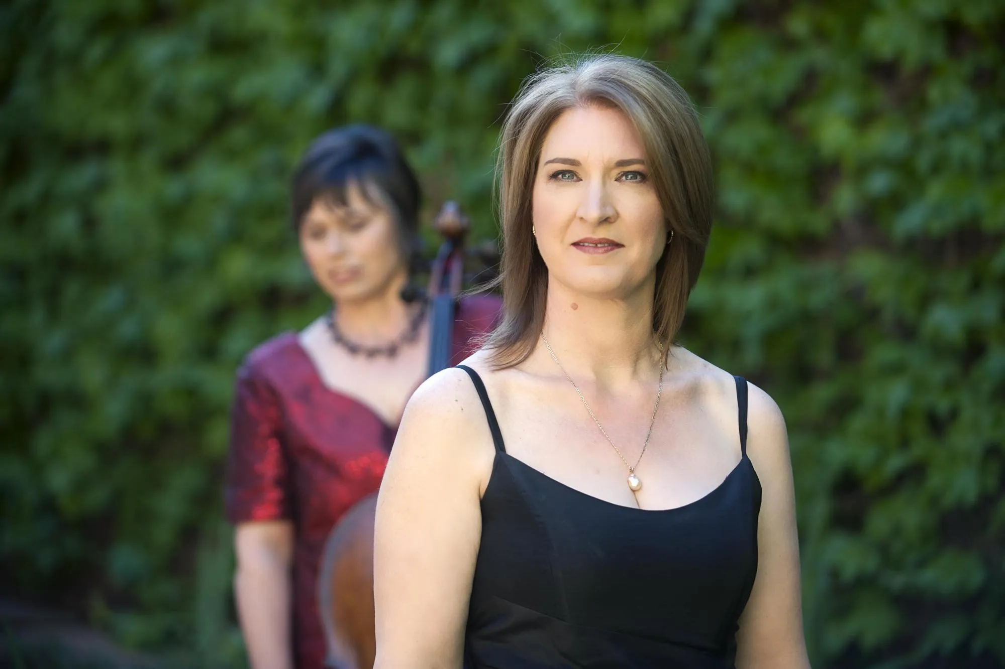 Ilse in focus, looking directly at the camera, with Anmari blurred in the background, both in front of a wall of green foliage. Anmari is wearing a dark red sequined blouse, and Ilse a black dress.