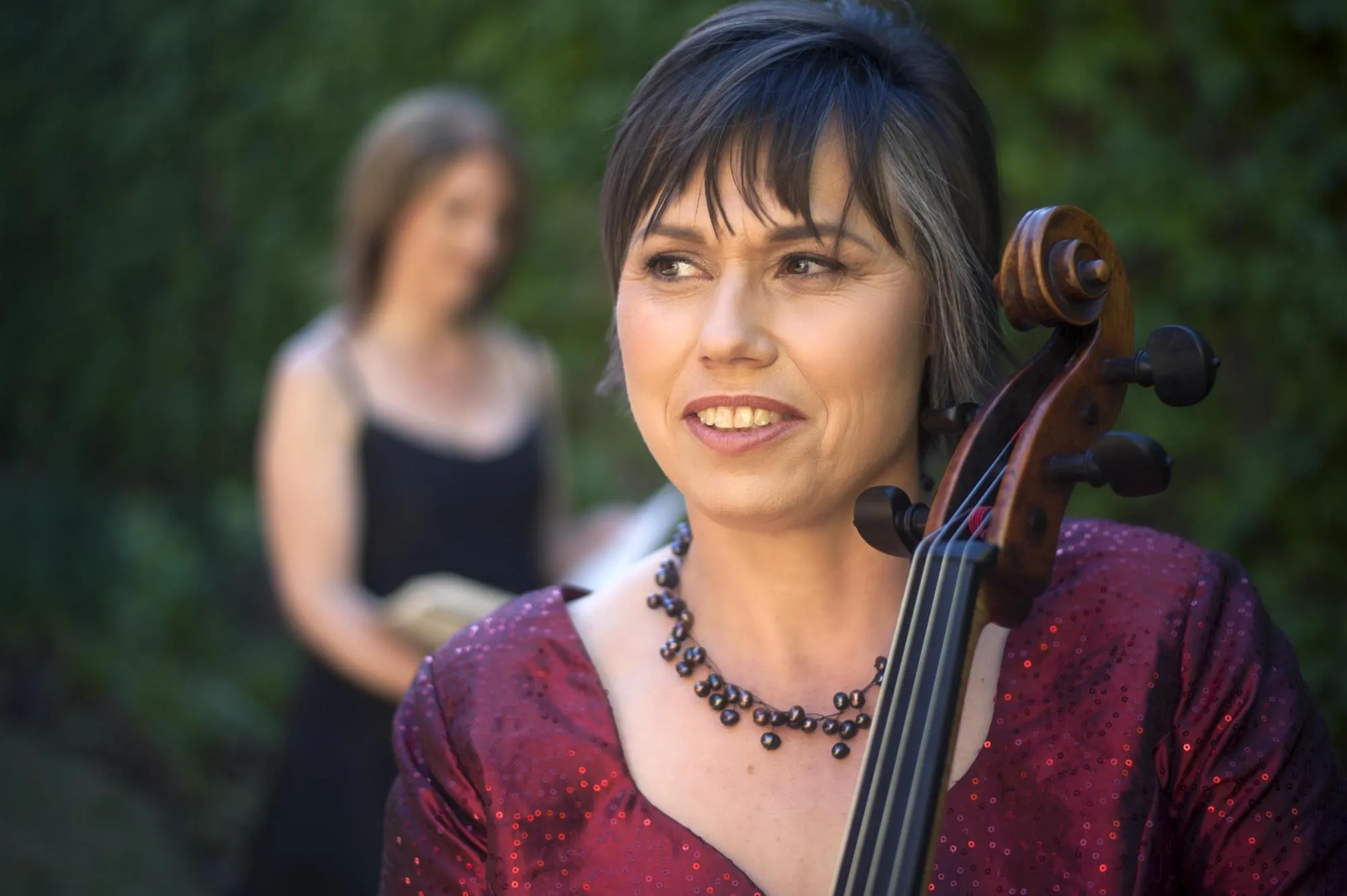 Anmari standing close to the camera, holding her cello, looking off into the distance. Ilse is blurred in the background, looking at a volume of sheet music. Both are in front of a wall of green foliage. Anmari is wearing a dark red sequined blouse, and Ilse a black dress.